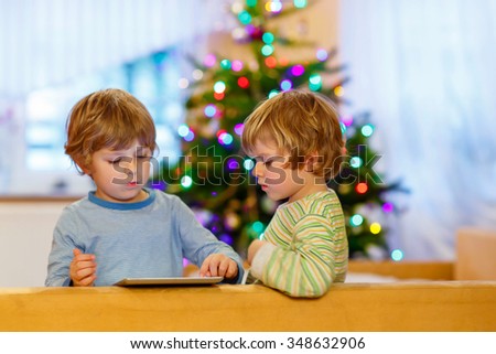 Two adorable little kid boys playing with tablet pc, indoors. Happy siblings toddlers, blond twins, looking at gift. With Christmas tree and lights on background.