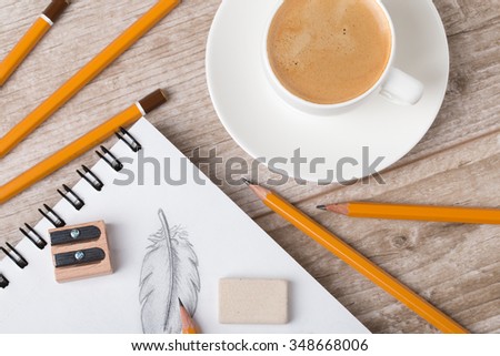 Close-up view of artist's or designer's table. Cup of coffee, pencils, sharpner and eraser laying on sketch book with hand-drawn feathers