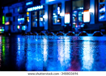 Rainy night in the big city, shop windows glowing in the reflection of asphalt. View from the level of asphalt, in blue tones
