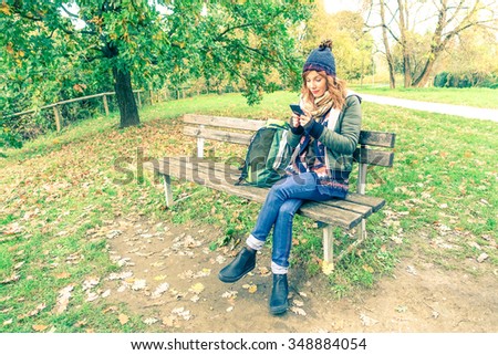 Young woman with red hair holding mobile phone sitting in a park bench wearing winter clothes and cap - Student girl with smartphone texting message outdoors in a green nature - Concept of technology