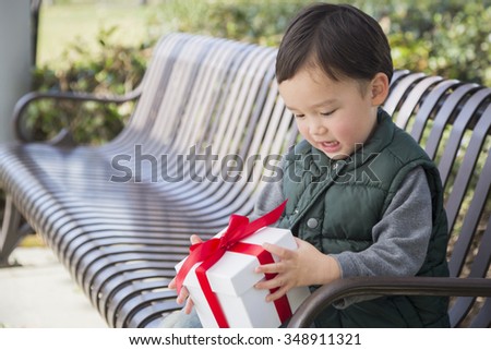 Adorable Mixed Race Boy Opening A Christmas Gift Outdoors On A Bench.