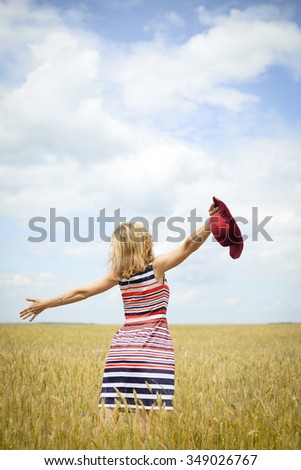 Picture of beautiful young woman wearing striped dress holding red hat on field. Backview of pretty girl excited on summer sunny farm field countryside background.