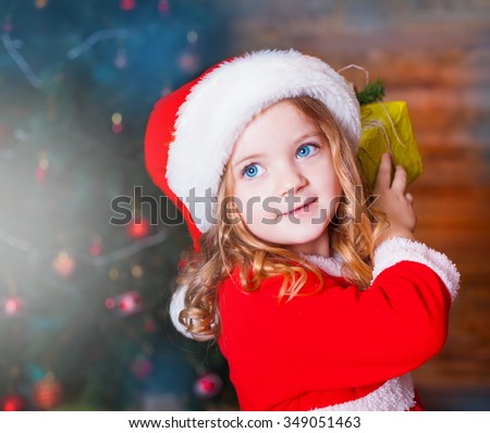 Beautiful little curly blonde girl, has happy fun cheerful smiling face, blue eyes, red Christmas hat, holding a gift box. Portrait holiday. 