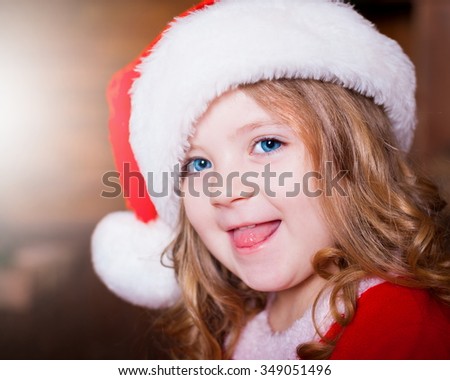 Beautiful little curly blonde girl, has happy fun cheerful smiling face, blue eyes, red Christmas hat. Portrait holiday. 
