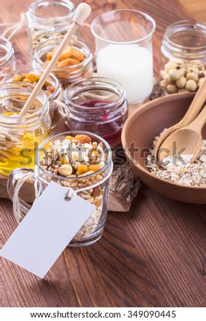Healthy morning muesli ingredients in glass cup with a blank tag for recipe. Raw rolled oats, dried fruits, seeds, honey, nuts and milk on rustic wooden board