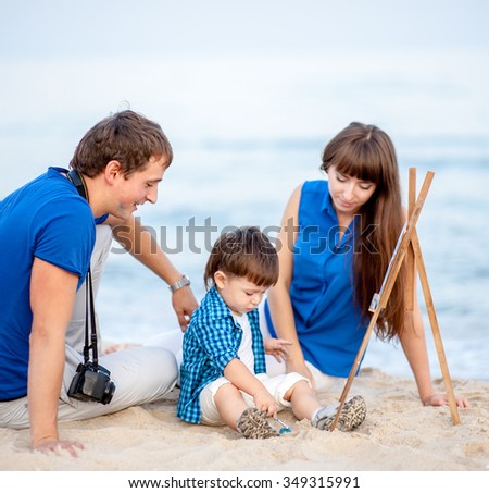 Woman, man and child in blue and white dress sit on the beach and draw on the easel near the surf