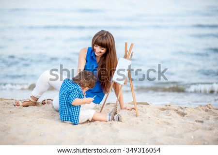 Woman and child in blue and white dress sit on the beach and draw on the easel