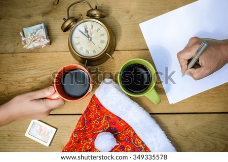 Top view picture of male hand writing on paper and female hand holding cup of coffee. Closeup of planning with alarm clock and Santa hat on wooden plank background.