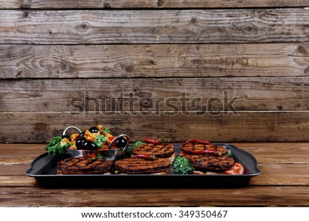 fresh red meat beef hamburger served on black tray with vegetables olives hot chili pepper tomatoes mushrooms over wooden table with empty space for text on background