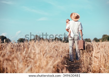 Picture of father holding baby and old suitcase in wheat field. Backview of happy family walking on summer countryside landscape background.
