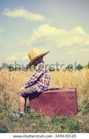 Picture of little boy in hat sitting on old suitcase in wheat field. Sideview of young traveller waiting on summer countryside background.