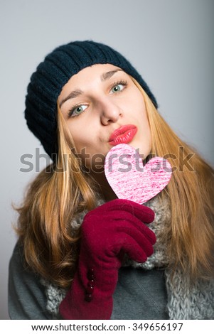 blonde girl holding a heart, symbol of love, dressed in winter clothing, Christmas concept, studio photo isolated on a gray background