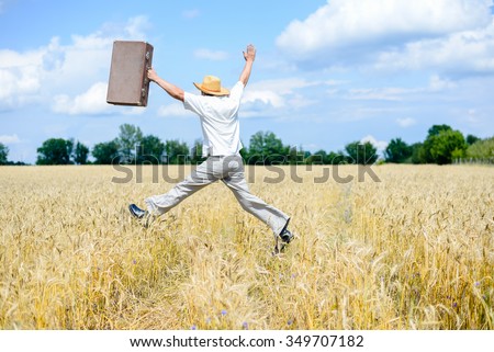 Picture of man in hat jumping with old suitcase on wheat field. Excited traveller on summer sunny countryside background.
