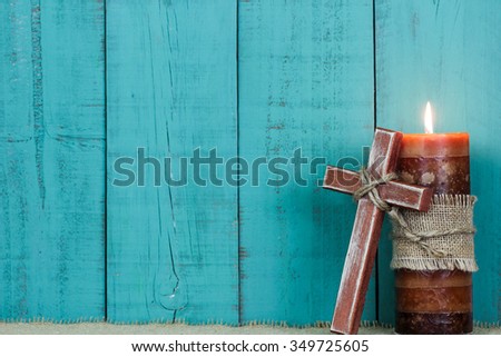 Textured candle burning and rugged wooden cross with rope by antique rustic teal blue wooden background; Easter, Christmas, Memorial Day and religious background with painted copy space