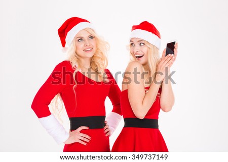 Funny playful sisters twins in red santa claus dresses and hats using one smartphone over white background
