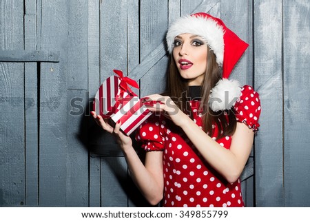 Closeup view portrait of one beautiful brunette young surprised new year woman celebrating christmas in red santa claus hat with white fur and dress holding present box indoor, horizontal picture