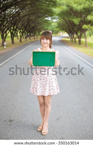 Cute woman hand holding green blank board sign on road with tree, Smiling female model.