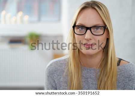 Serious thoughtful young woman with long blond hair wearing glasses looking pensively at the camera, blurred indoor home background with copy-space