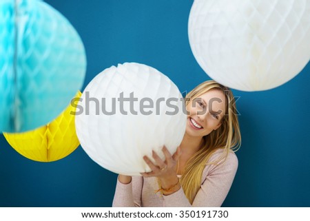 Happy attractive young blond woman celebrating Christmas and the festive season holding a big paper lattice ball decoration in her hands grinning around the side at the camera, over blue