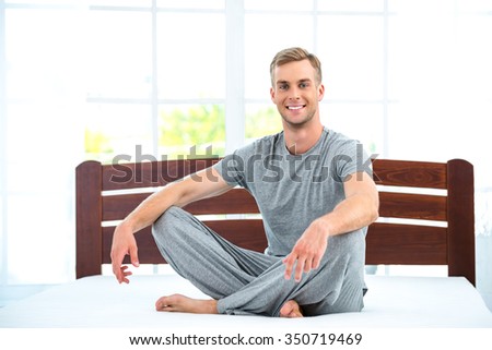 Photo of young man sitting on nice white bed. Young man demonstrating quality of mattress