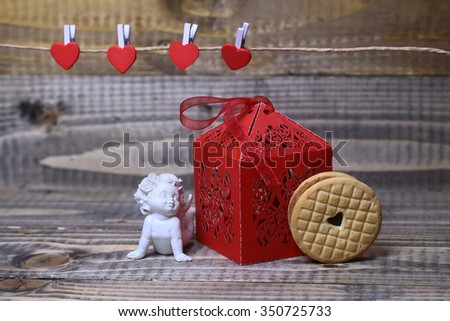 Closeup view of beautiful cupid angel decorative figurine near red paper greeting valentine box near clothes-peg in shape of heart with round pastry on wooden background copy space, horizontal picture