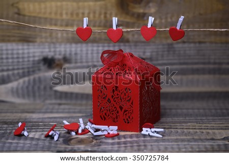 Closeup view of one beautiful decorative red paper greeting valentine box with ribbon bow near hanging clothes-peg in shape of heart with no people on wooden background, horizontal picture