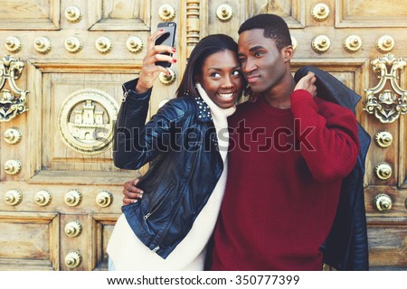 Two smiling best friends making self portrait on smart phone camera while standing against retro door, beautiful stylish couple photographing themselves on cell telephone for social network picture 