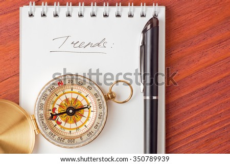 Closeup of notepad with handwritten word Trends, compass and pen on wooden board top view have place for text