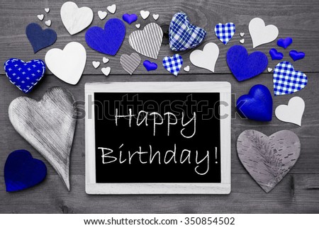 Chalkboard With English Text Happy Birthday. Many Blue Textile Hearts. Wooden Background With Vintage, Rustic Or Retro Style. Black And White Image With Colored Hot Spots.