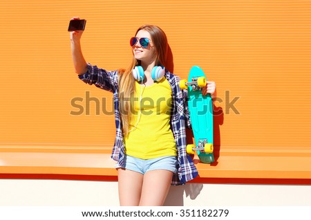 Pretty cool smiling girl in sunglasses with skateboard taking picture self portrait on smartphone over orange background