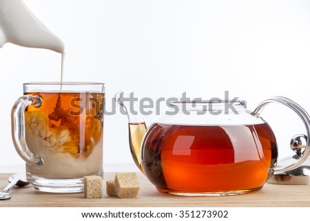 Dissolve milk in a cup of black tea. Transparent teapot and cup with three cubes of brown sugar on white background