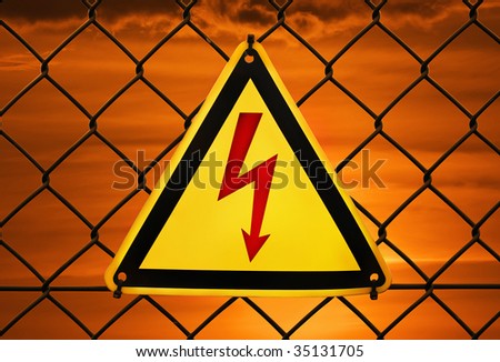 Metal warning sign "High voltage" tied to a fence with dramatic sunset sky on background.