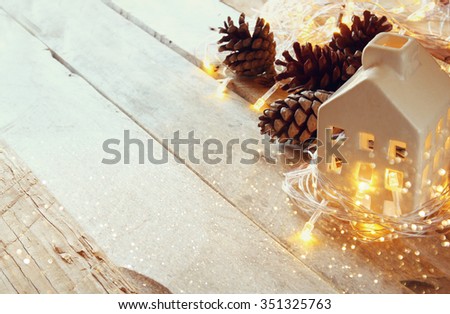 photo of pine cones and decorative wooden house next to gold garland lights on wooden background. copy space. retro filtered
