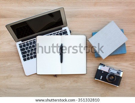 Laptop, film camera, Notepad with pen and a couple of books on the wooden table.