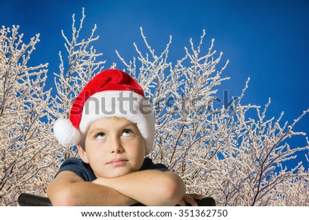 The charming seven-year-old boy in red Santa Claus cap thoughtfully looks up. The picture is made against the snow-covered New Year's wood