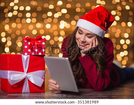 Thoughtful smiling young woman in santa claus hat lying on the floor and using tablet over sparkling background