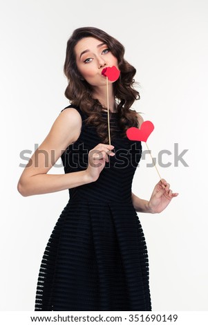 Amusing charming retro styled curly woman in black dress posing with paper heart and lips booth isolated over white background