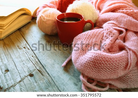 selective focus photo of pink cozy knitted scarf with to cup of coffee, wool yarn balls  and open book on a wooden table. style retro filtered and toned

