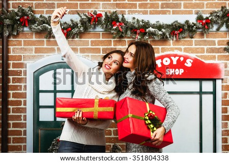 Two young attractive woman holds presets boxes in hands and making selfie on christmas street backdrop