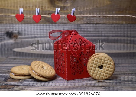 Closeup view of beautiful decorative red paper greeting valentine box with ribbon near clothes-peg in shape of heart with round pastry on wooden background, horizontal picture