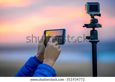 Female hands holding phone near the camera on the tripod. Remote control action camera filming time lapse video in the early morning