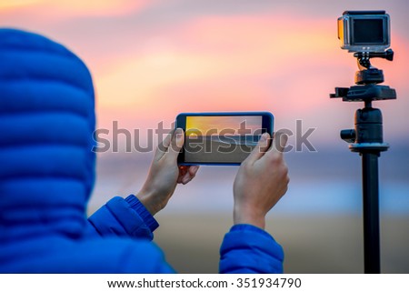 Female hands holding phone near the camera on the tripod. Remote control action camera filming time lapse video in the early morning