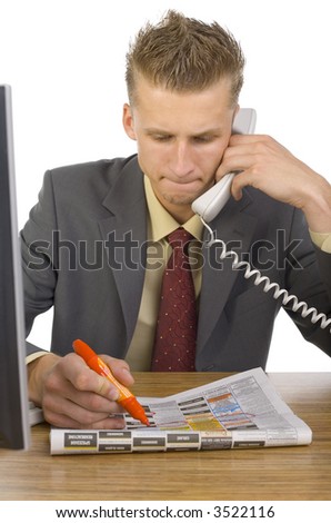 Young businessman sitting at desk, talking on phone and marking something in newspaper. White background, front view