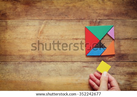 man's hand holding a missing piece in a square tangram puzzle, over wooden table.