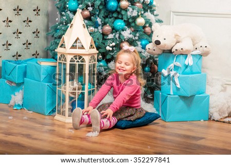 Little pretty curly blonde smiling girl sitting nearly Christmas tree with Christmas decorations and presents
