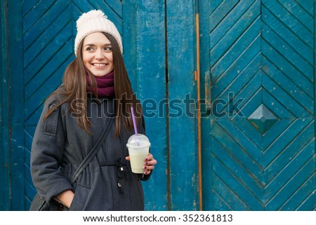 Pretty smiling girl, wearing in gray jacket and burgundy scarf, posing with cup of smoothie with banana and kiwi, with blue wooden doors on the background, waist up