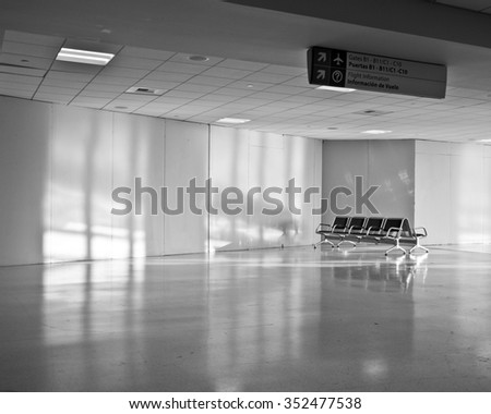 empty seats in large open deserted airport terminal with signs for gates on ceiling and shadows from cast onto the blank walls. black and white