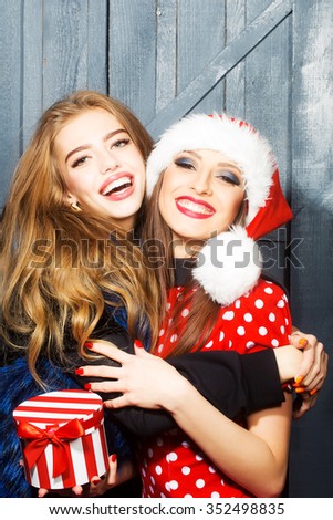 Closeup view of two beautiful brunette young happy smiling embracing new year women celebrating christmas in red santa claus hat with white fur holding gift box indoor, horizontal picture