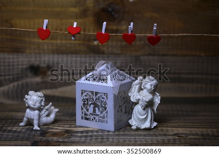 Closeup view of two beautiful cupid angels decorative figurine near white paper greeting valentine box near red clothes-peg in shape of heart with no people on wooden background, horizontal picture