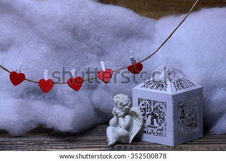 Closeup view of one beautiful cupid angel decorative figurine near paper greeting valentine box and hanging red clothes-peg in shape of heart with white wadding decorating snow, horizontal picture